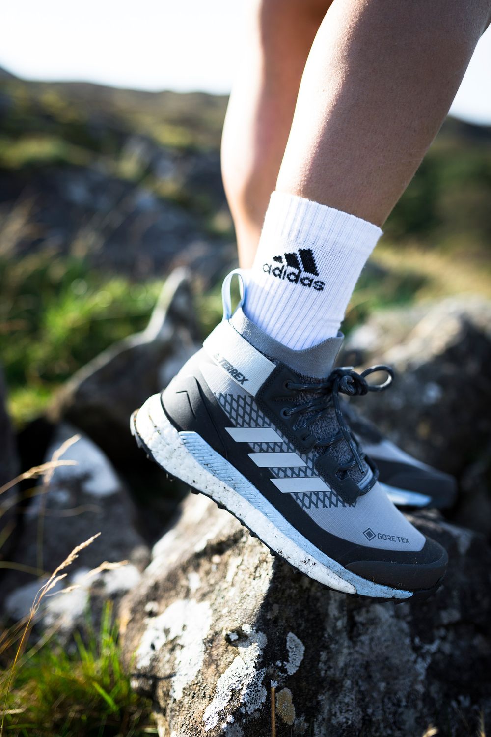 Person wearing Adidas GORE-TEX trail running shoes with white Adidas socks, stepping on rocky terrain outdoors.