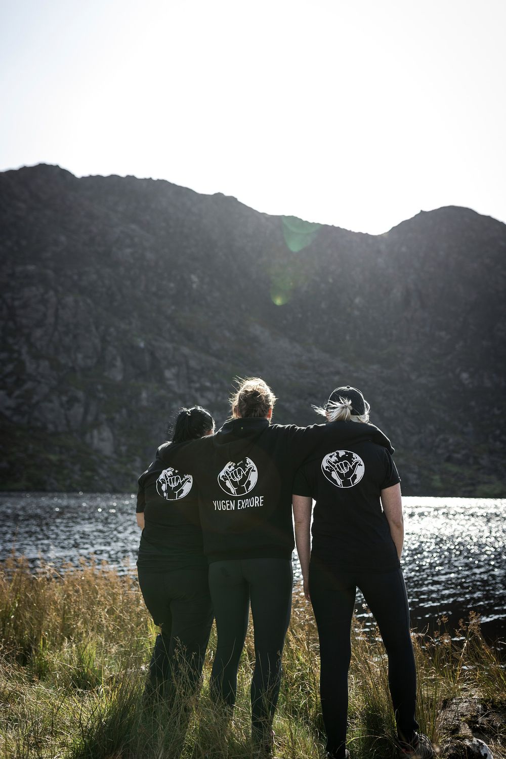 Three friends in matching black t-shirts stand together facing a mountain lake in a scenic outdoor landscape.