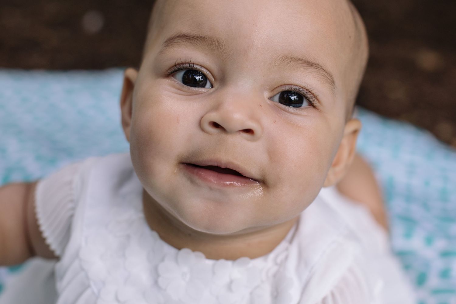 A smiling woman in an orange top holds a happy baby outdoors with lush green bushes in the background.