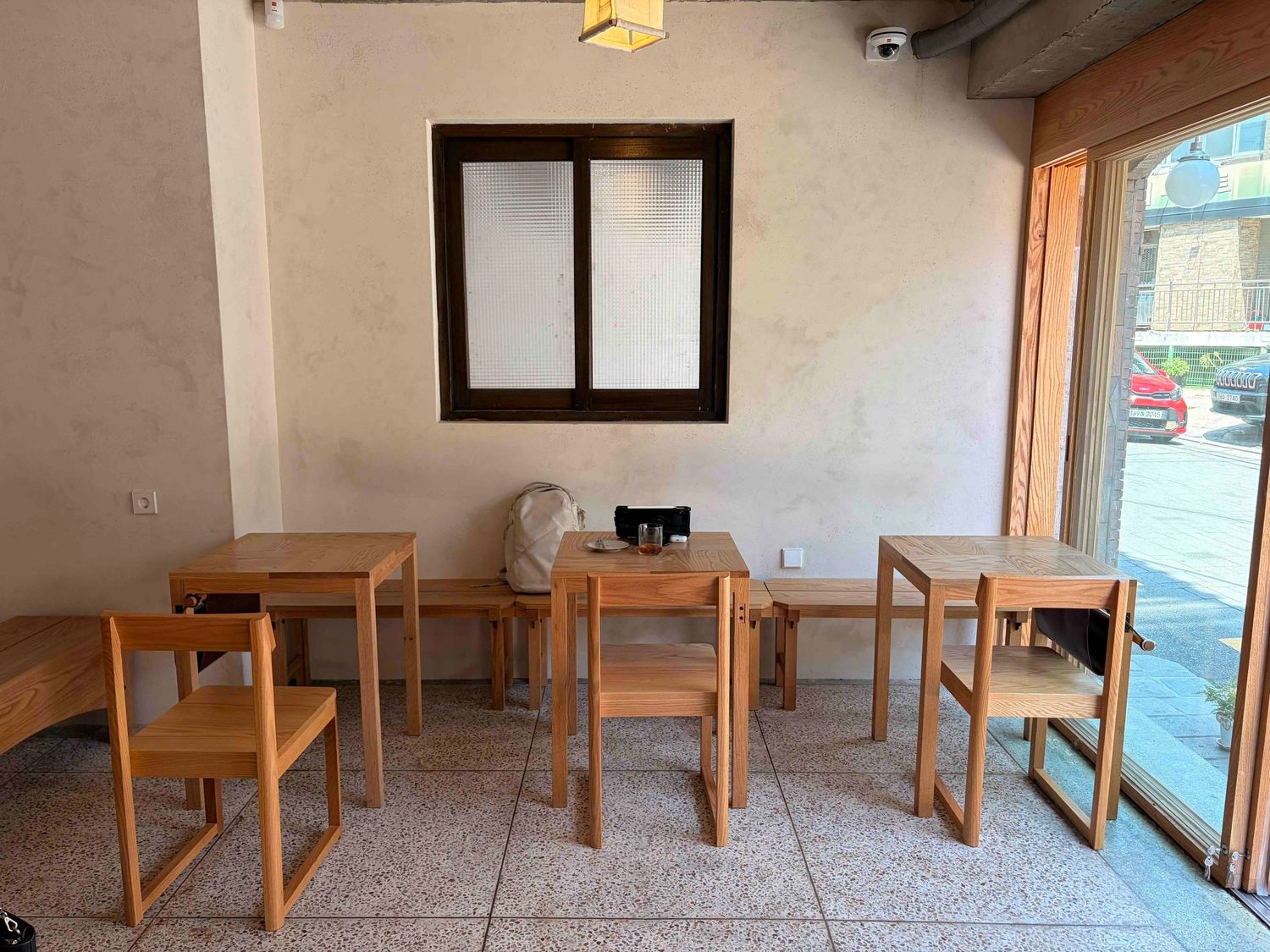 Minimalist cafe interior with wooden tables and stools on tiled floor, white walls, and a framed window.