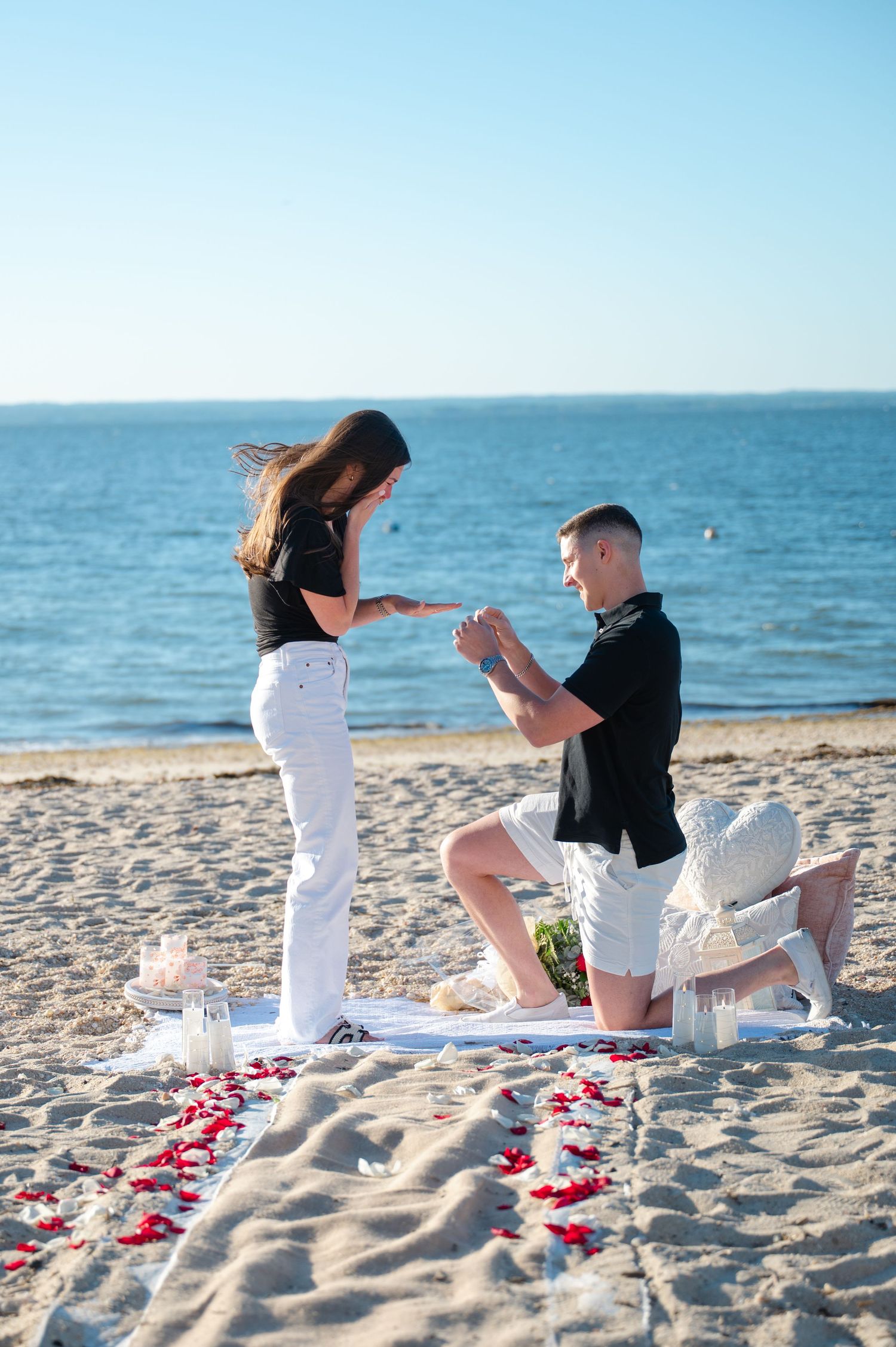 Man proposes to woman on sandy beach with rose petals and candles, ocean in background on sunny day.