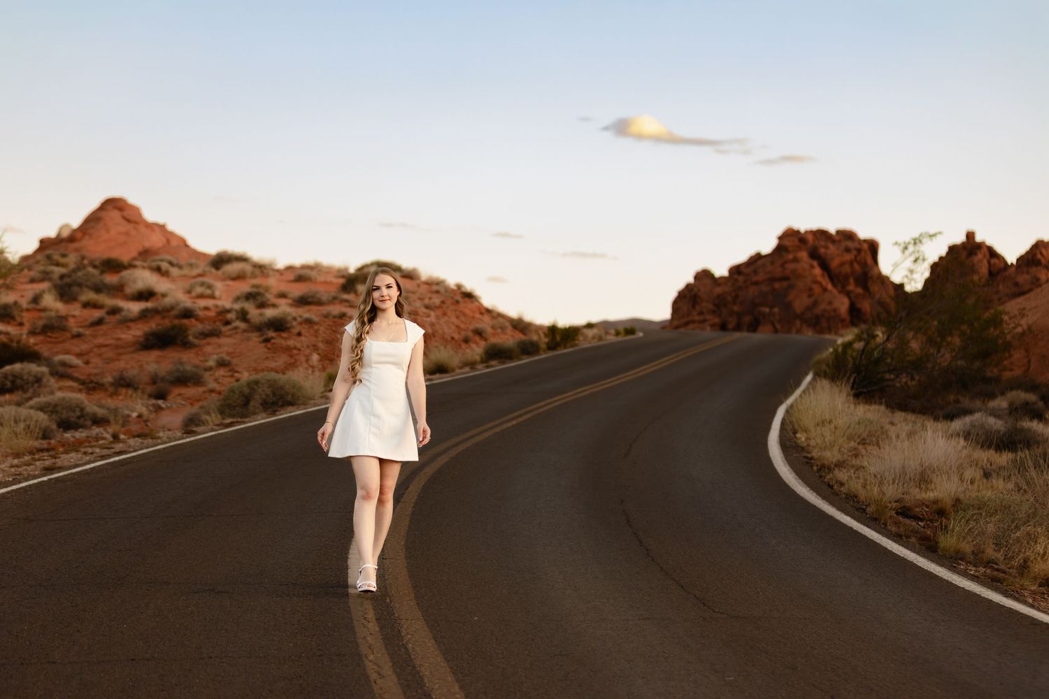 Young woman in white dress walking on winding desert road surrounded by red rock formations at golden hour.