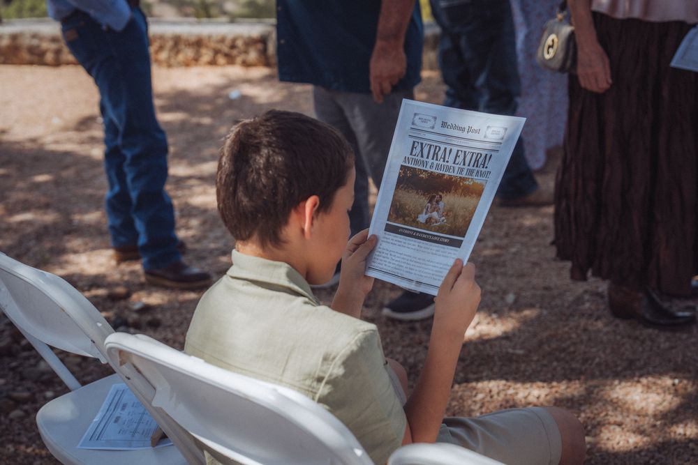 Young boy reading an Extra edition newspaper outdoors at a gathering, seated in a white folding chair.