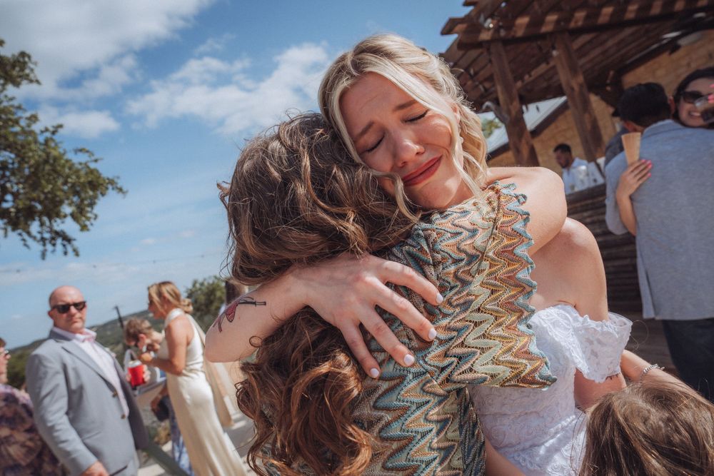 Bride in white lace dress sharing a warm embrace with a guest in a patterned dress at outdoor wedding.