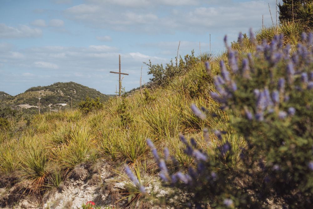 Wooden cross on a hillside surrounded by lush green grass and purple wildflowers under a partly cloudy sky.