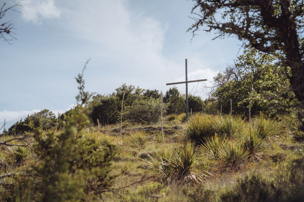 A wooden cross stands atop a lush green hill surrounded by native shrubs and trees under a cloudy sky.