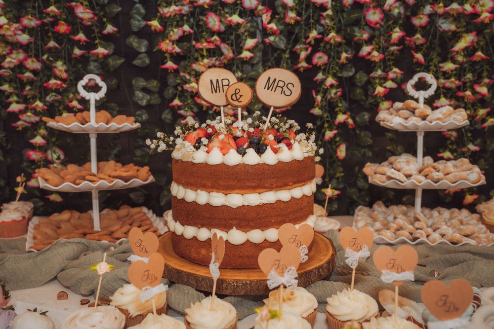 Rustic wedding dessert table featuring a naked Mr & Mrs cake with white frosting, cupcakes, and assorted sweets against a floral backdrop.