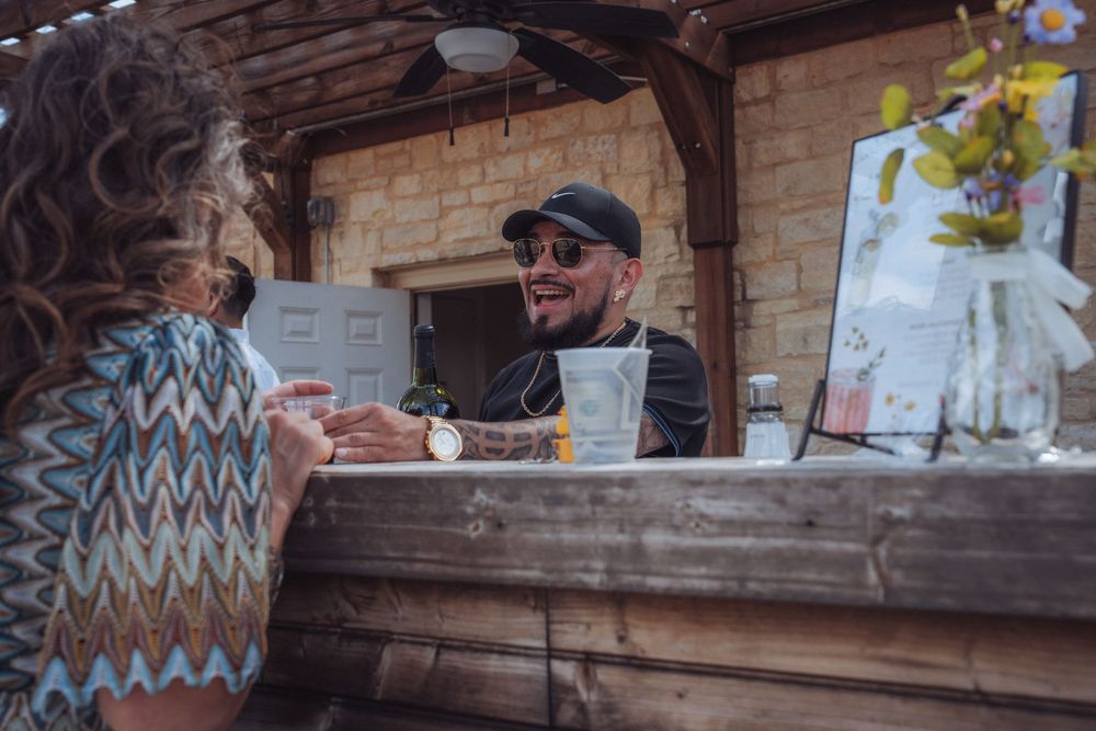 Smiling man with sunglasses and cap serving customer at outdoor wooden bar counter.