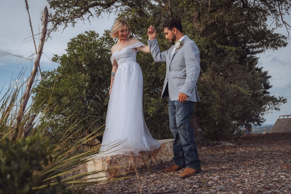 Bride in white gown being twirled by groom in gray blazer and jeans among trees at outdoor wedding.
