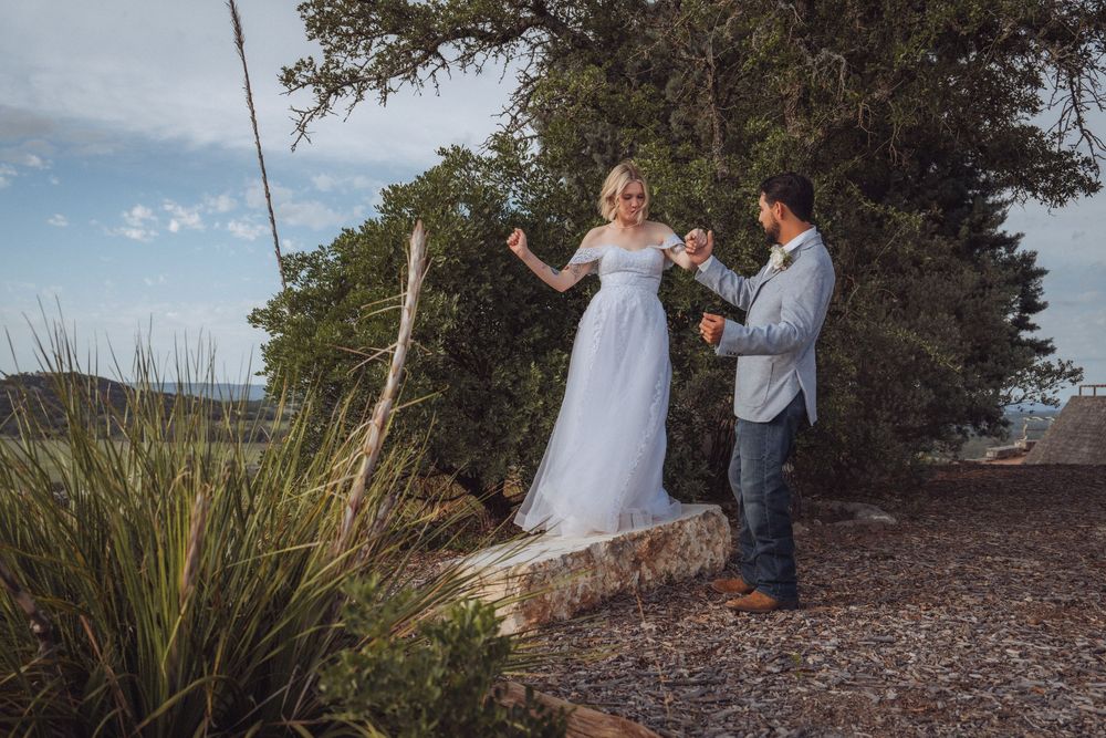 Bride in white gown and groom share a joyful moment outdoors near rustic wooden swing at scenic wedding venue.