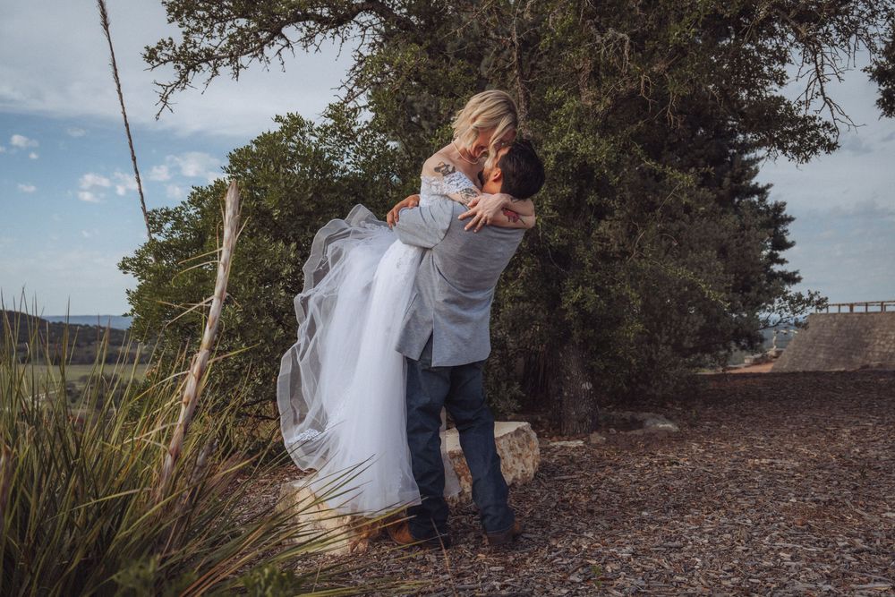 A groom lifts his bride in a white wedding gown outdoors near trees in a scenic countryside setting.