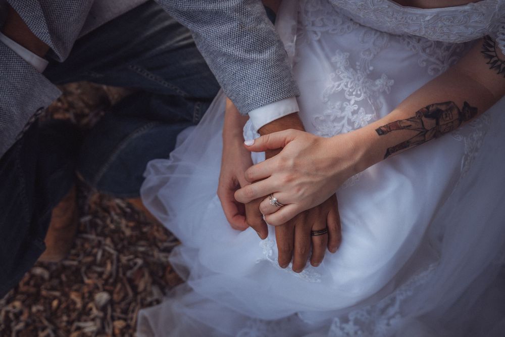 Close-up of bride and groom holding hands, showcasing engagement ring and floral tattoo on wedding day.