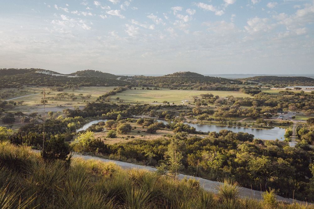 Aerial view of a winding road through lush green Texas Hill Country landscape with a river and rolling hills.