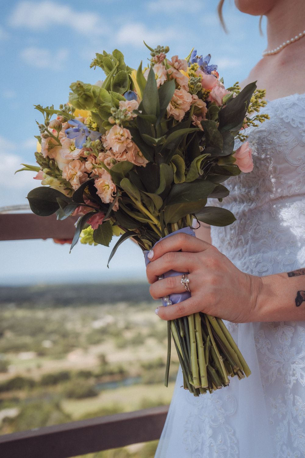 Bride in white lace dress holding a colorful wedding bouquet with pink, blue, and yellow flowers outdoors.