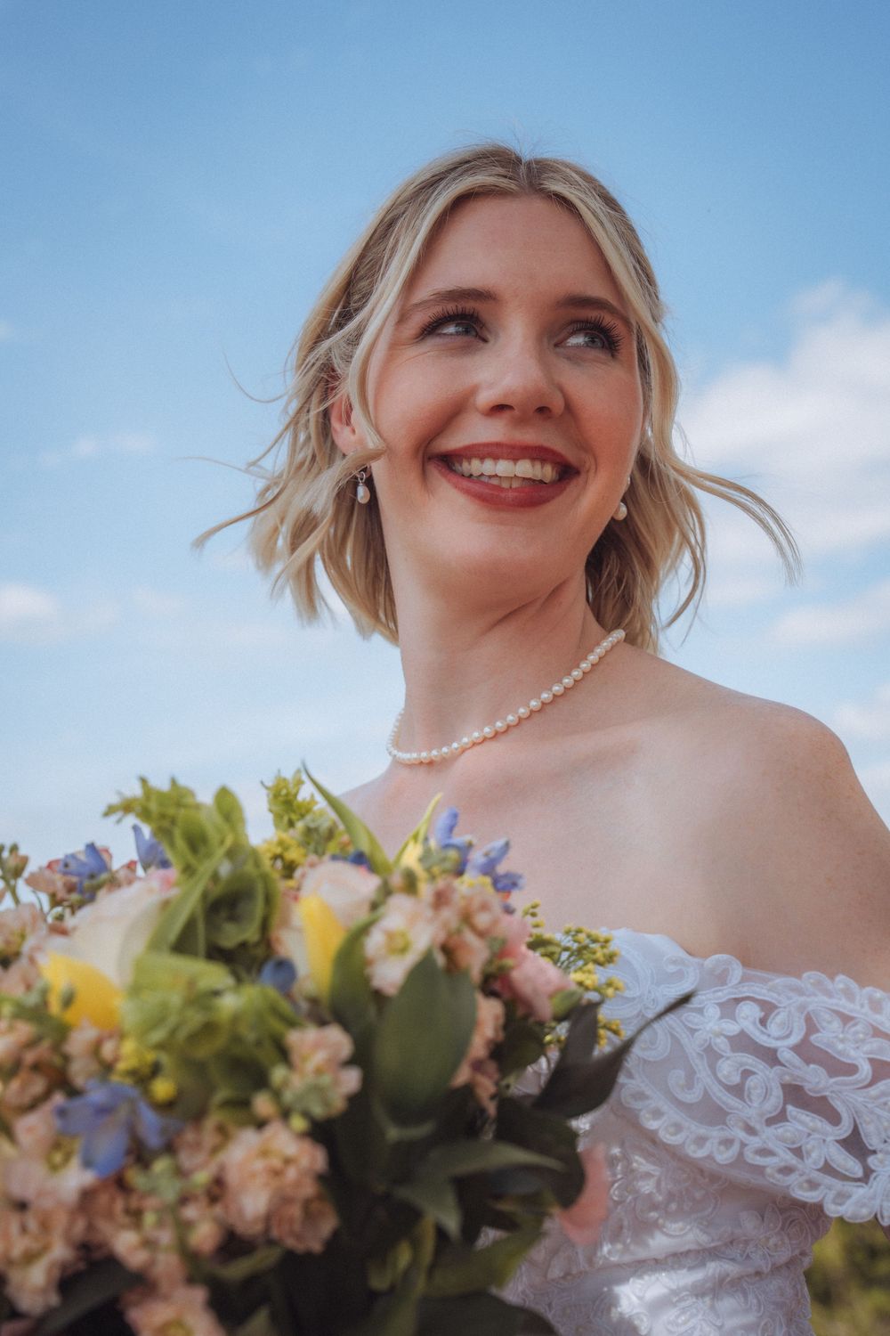 Smiling blonde bride in off-shoulder lace gown wearing pearl necklace holding a colorful bouquet under blue sky.