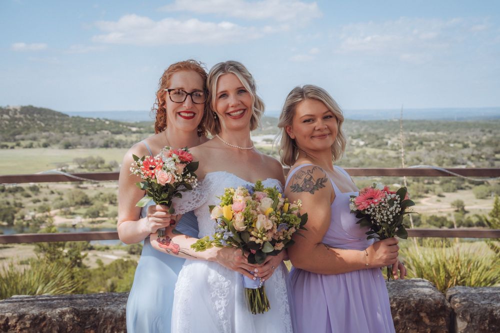 Bride in white gown poses with two bridesmaids in blue dresses holding bouquets with scenic landscape backdrop.