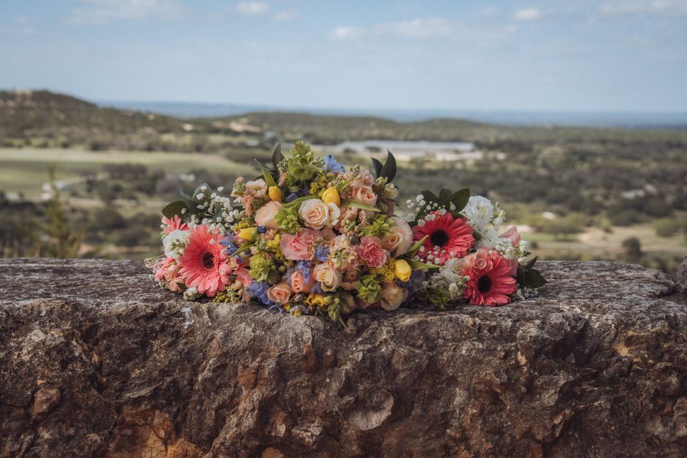 Colorful wedding bouquet with pink gerberas and wildflowers resting on a rustic stone wall with scenic landscape views.