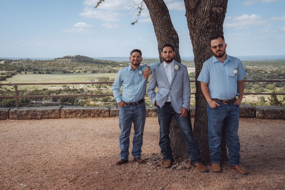 Groom in gray suit stands with two groomsmen in blue shirts and jeans beside a tree at an outdoor wedding venue.