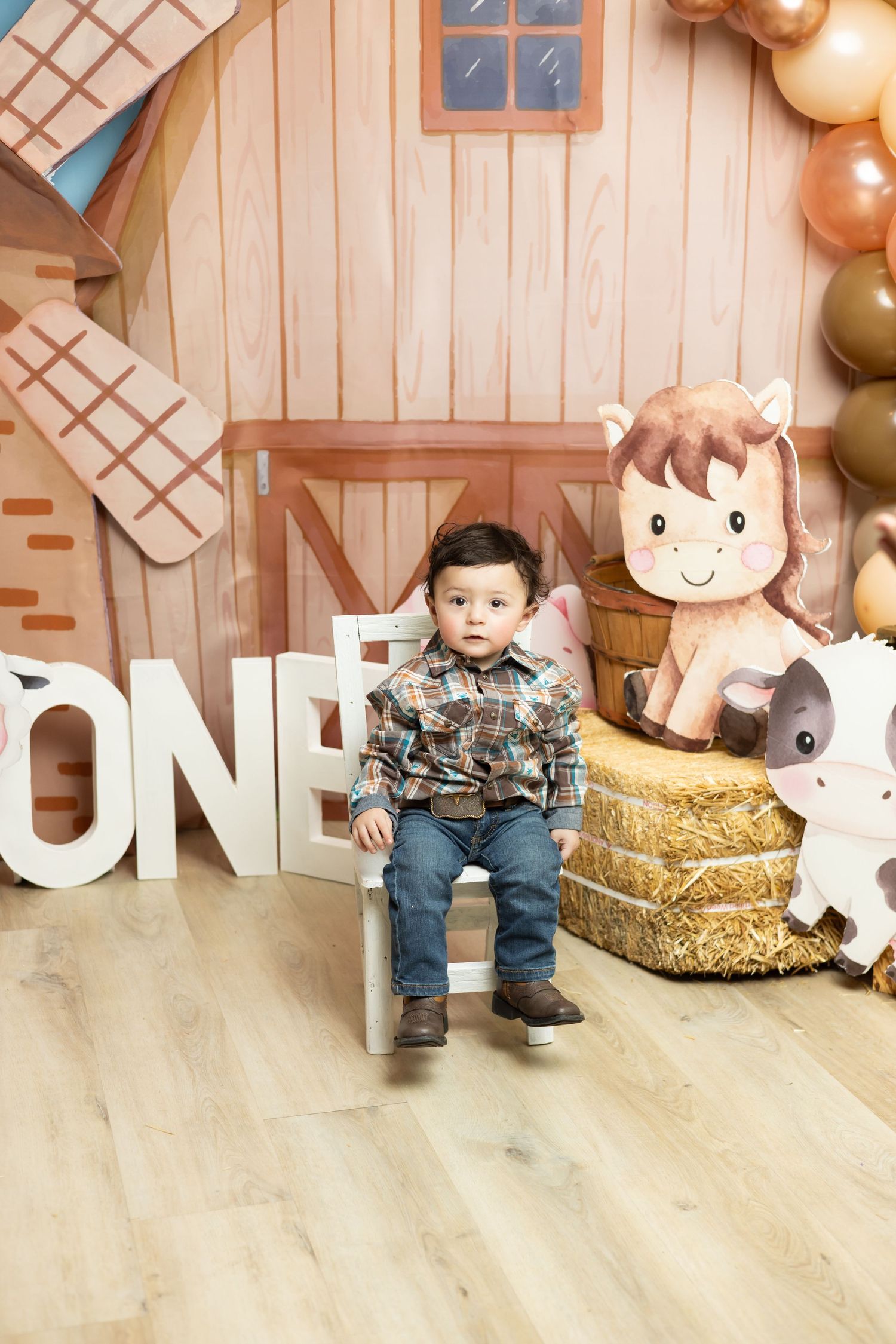 Toddler boy in plaid shirt sits on white chair at farm-themed first birthday party with ONE letters and hay bales.