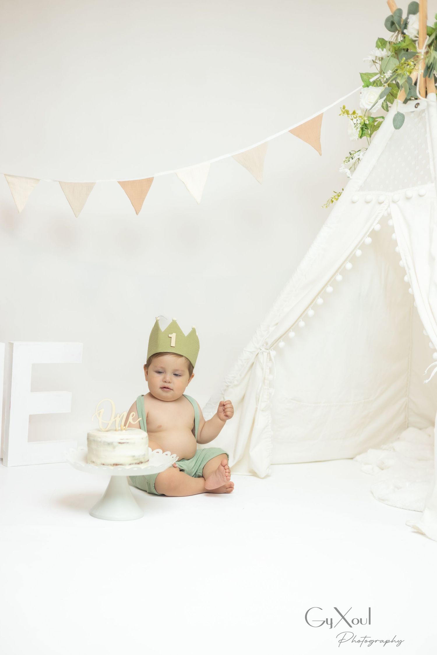 Baby's first birthday cake smash photo with green crown, white teepee, bunting, and small cake on white pedestal stand.