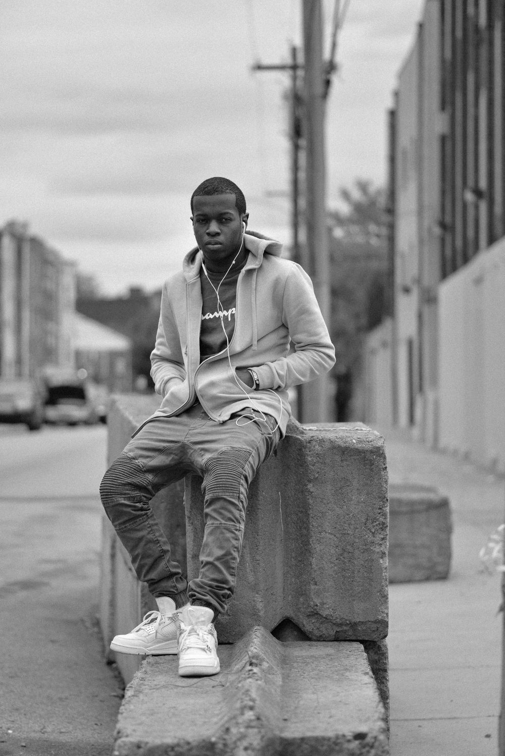 Young man in hoodie and sneakers sits on concrete barrier in urban alleyway, black and white photo.