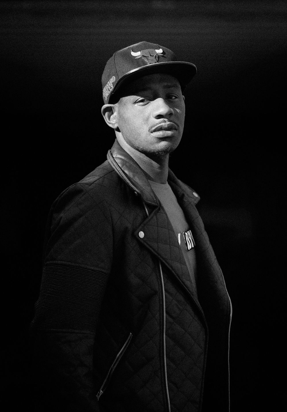 Black and white portrait of a young man wearing a Chicago Bulls cap and quilted jacket, looking over his shoulder.