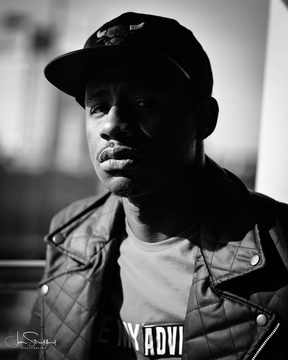 Black and white portrait of a young man wearing a snapback cap and quilted jacket outdoors.