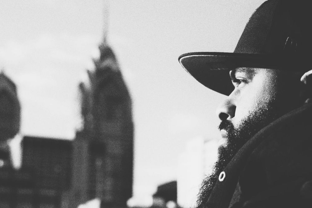 Bearded man in a snapback hat with city skyline in background, black and white street portrait.