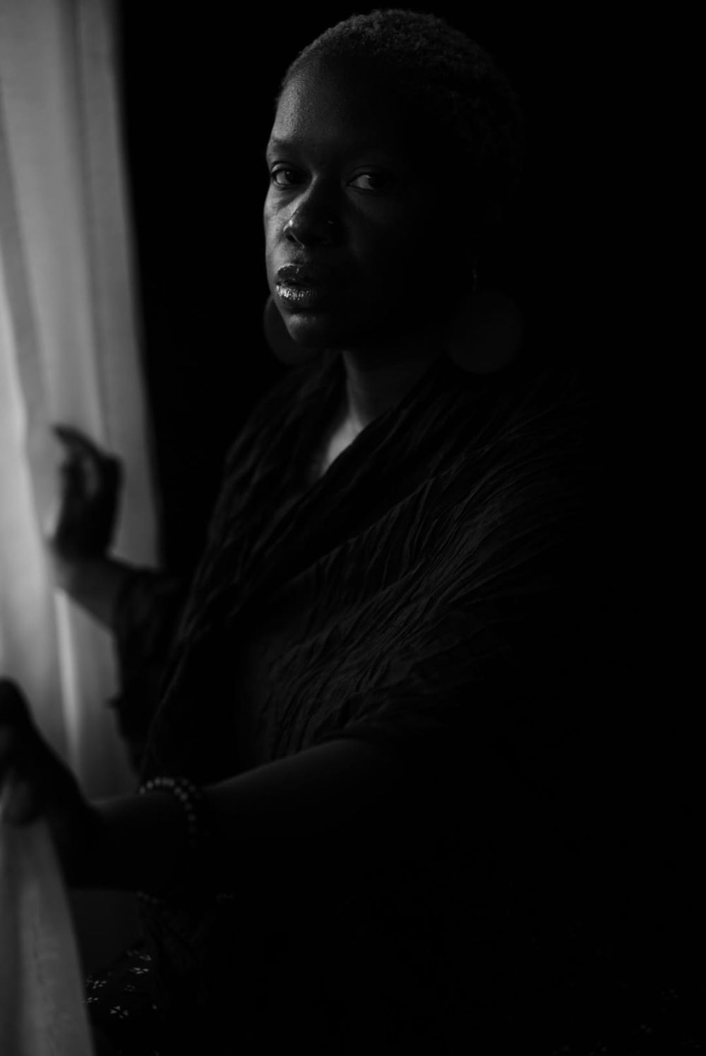 Black and white portrait of a woman standing by a window in dramatic low-key lighting.