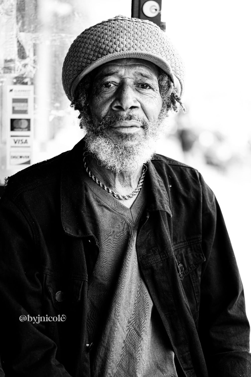 Black and white portrait of an elderly man wearing a knit cap and jacket, with a gray beard and chain necklace.