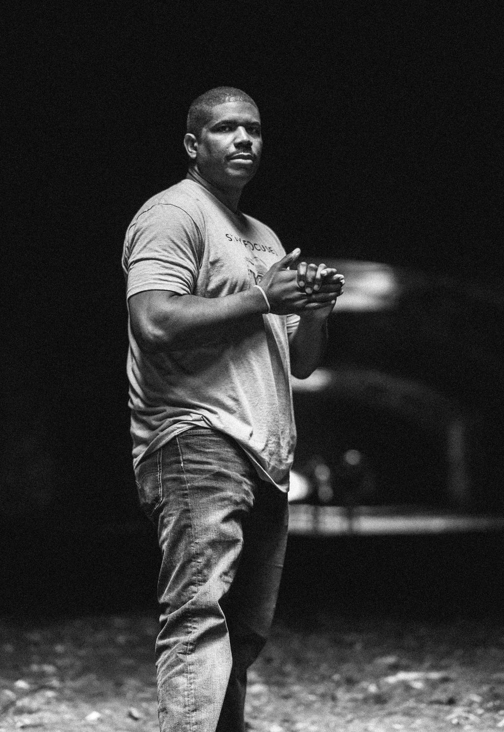 Black and white photo of a muscular man in jeans and t-shirt standing outdoors at night with hands clasped.