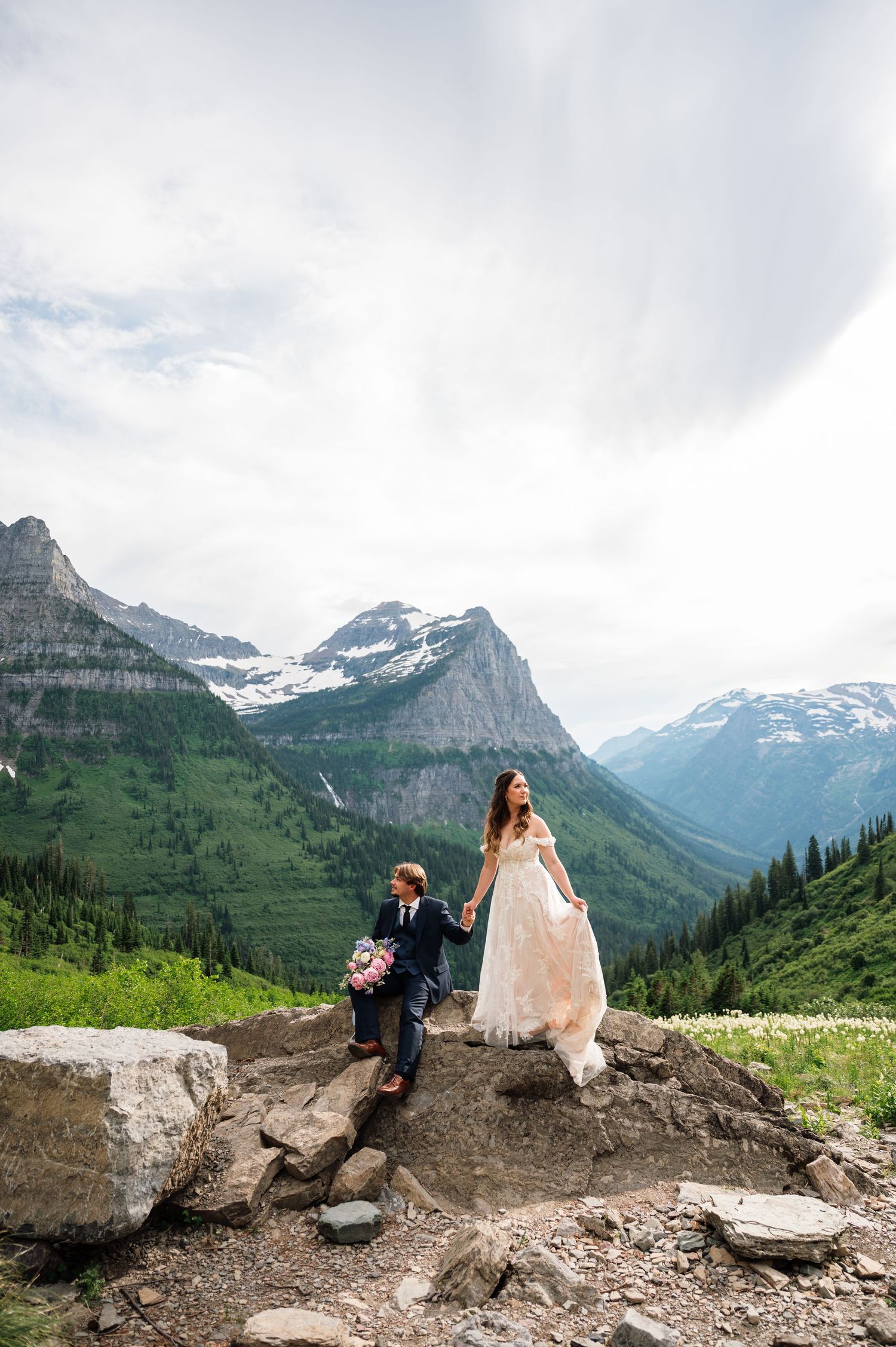Bride and groom hold hands on rocky mountain overlook with snow-capped peaks and green valleys in Glacier National Park.
