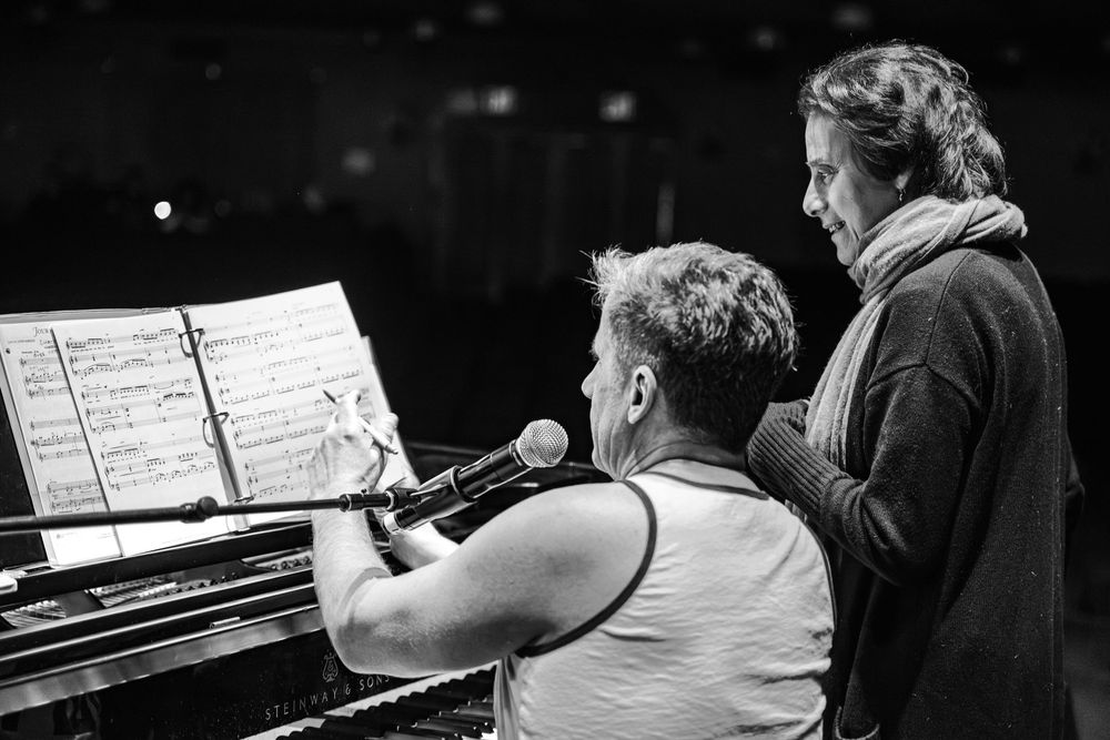 Two musicians collaborate at a piano, reviewing sheet music together in a black and white performance setting.