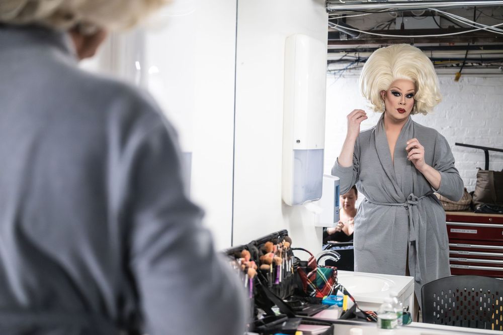 Drag performer in gray robe and blonde wig applying makeup while looking in a mirror at a backstage vanity.