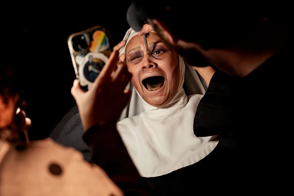 A distressed nun in costume screams while someone holds a phone nearby in a dramatic theatrical scene.