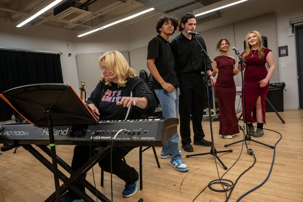 A musician plays Roland keyboard while four singers perform behind microphones in a rehearsal room.