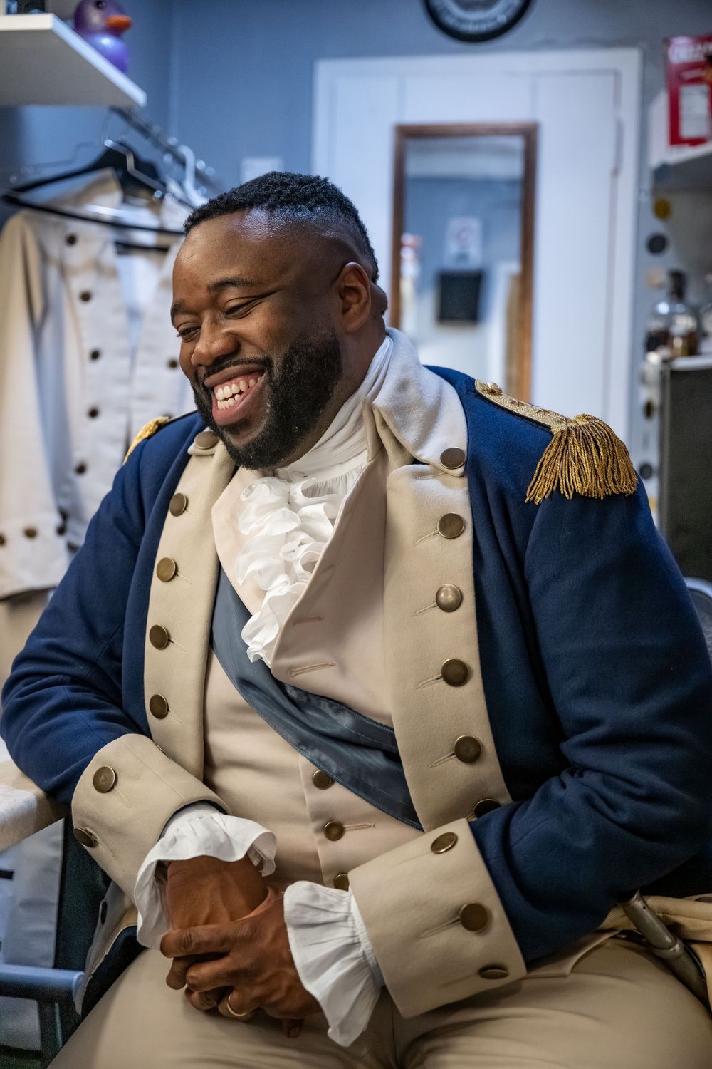 Man in colonial military costume laughing backstage, wearing navy blue coat with gold epaulettes and white ruffled shirt.