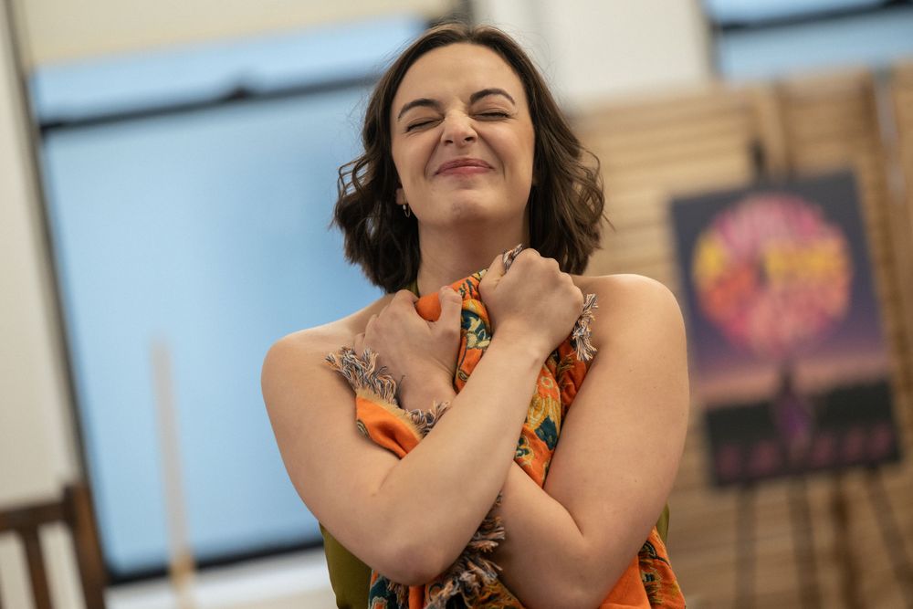Smiling woman with tattoos hugging herself, wearing a floral dress in a bright studio setting.