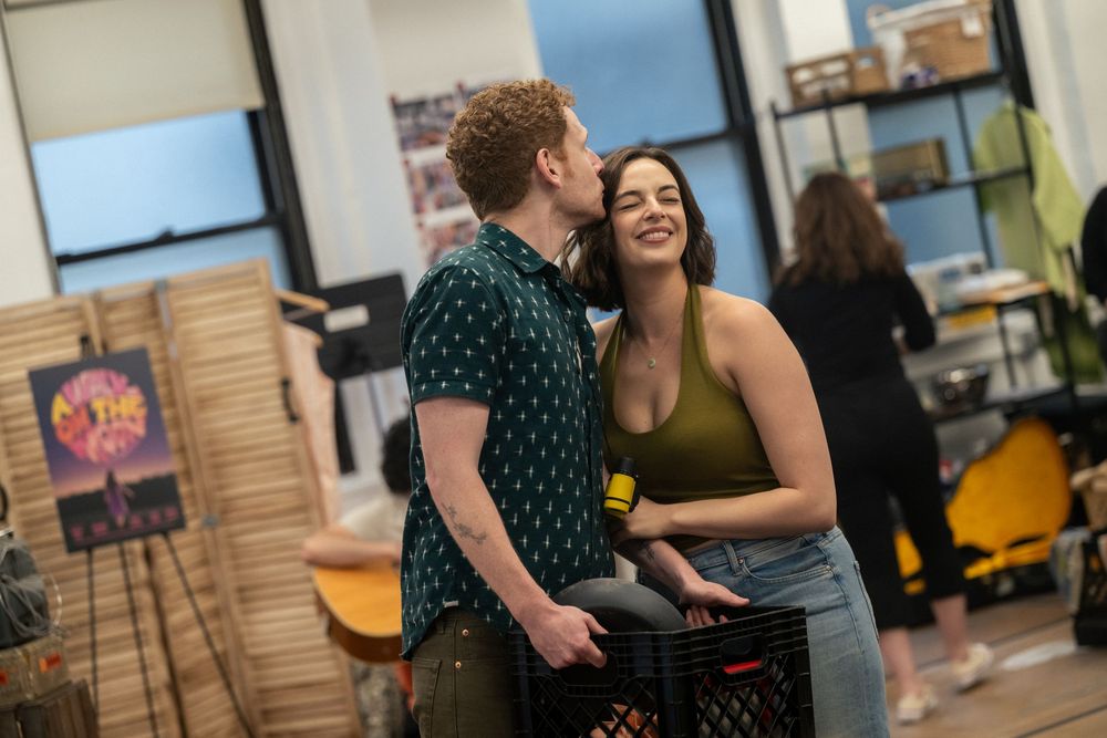 A man kisses a smiling woman on the cheek as she holds a black milk crate in a bright, casual indoor space.