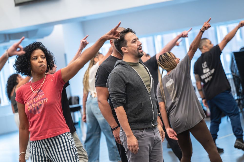 Diverse group of adults in a dance rehearsal, arms raised and pointing, practicing choreography in a bright studio.