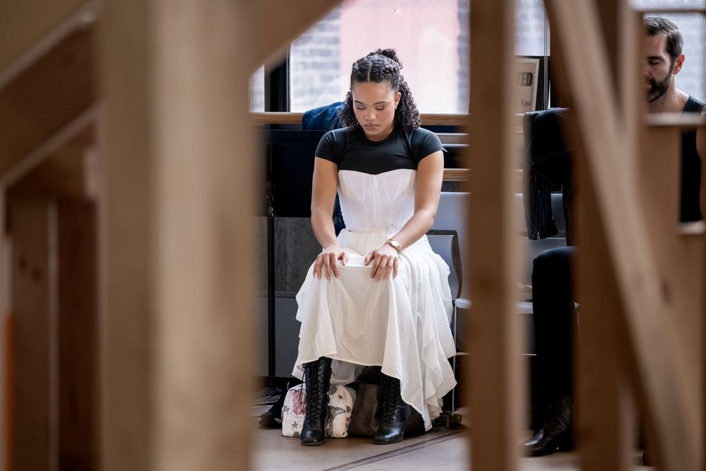 Woman in white dress and boots sits pensively in wooden church pew, head bowed in quiet reflection.