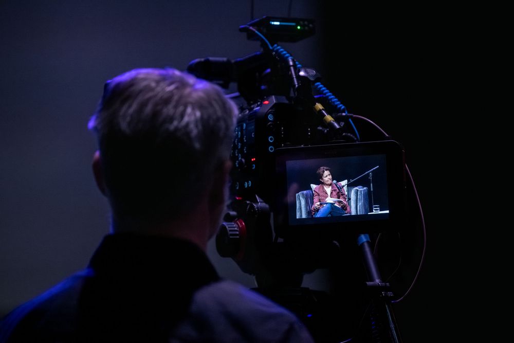 Cameraman filming a speaker on stage, visible in camera LCD screen during a live event production.