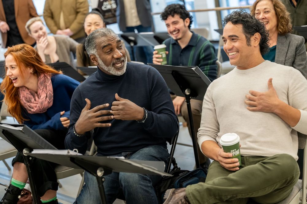 Two men laugh joyfully during a rehearsal, one holding a coffee cup, surrounded by smiling colleagues with music stands.