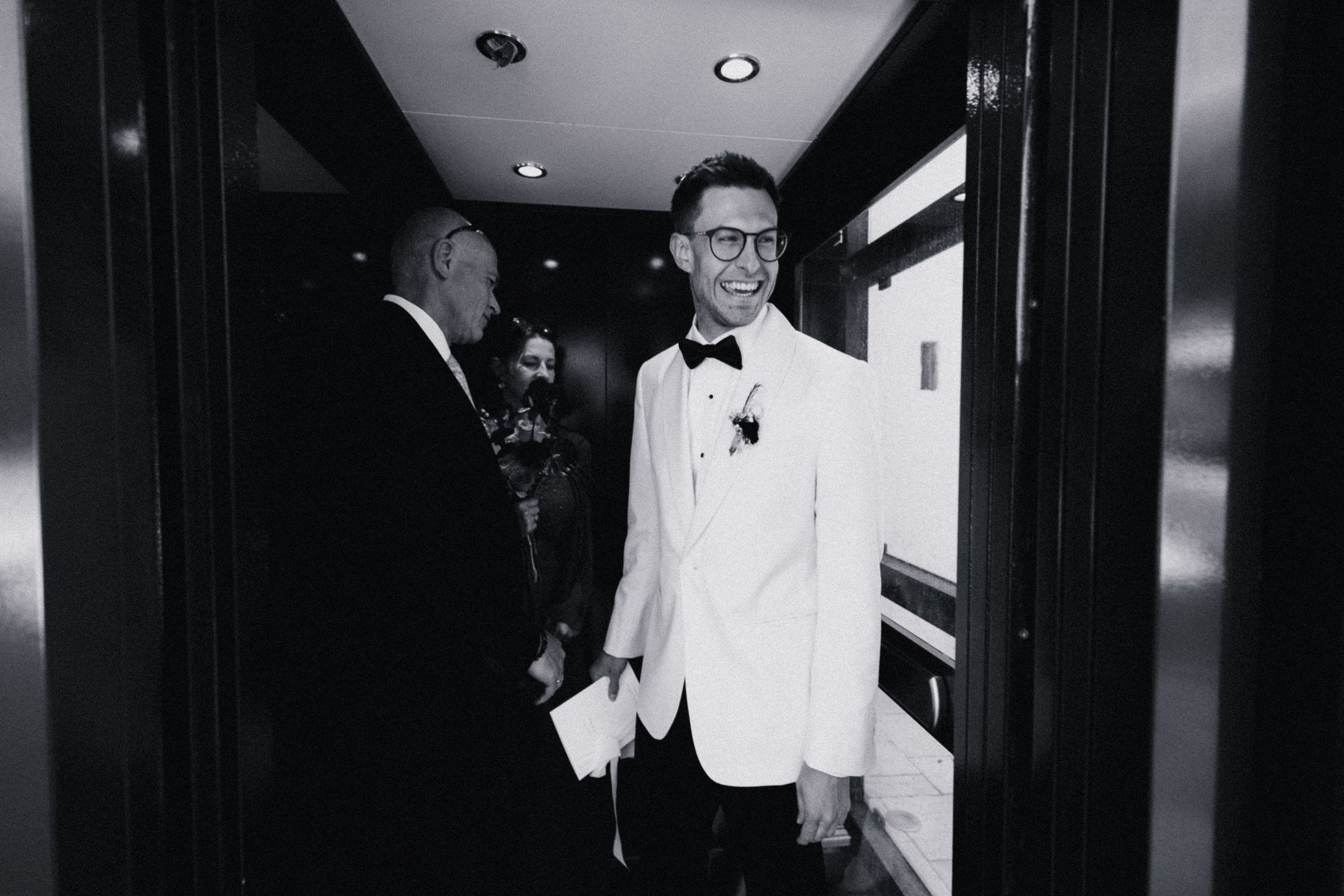 Groom in white tuxedo jacket smiling in hotel hallway during wedding day preparations, black and white photo.
