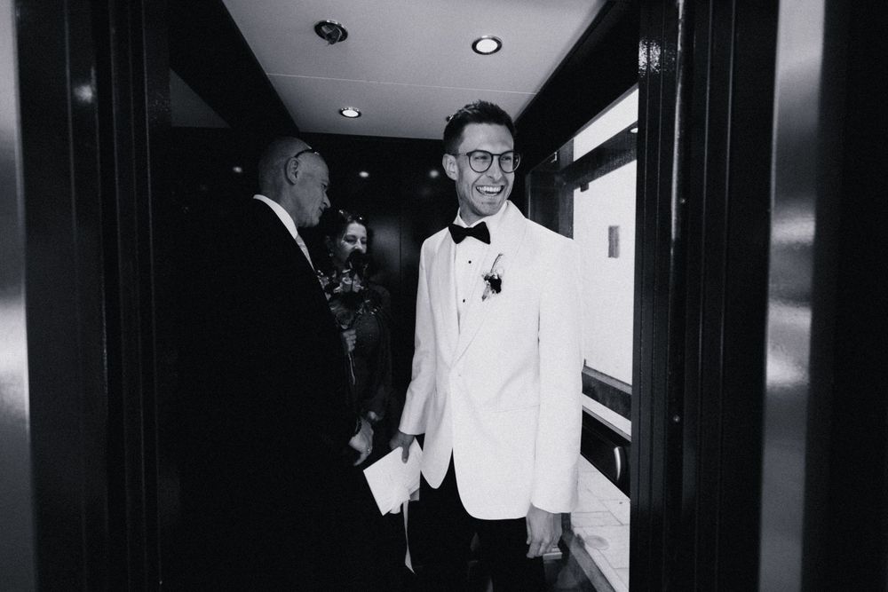 Groom in white tuxedo jacket smiling in hotel hallway during wedding day preparations, black and white photo.