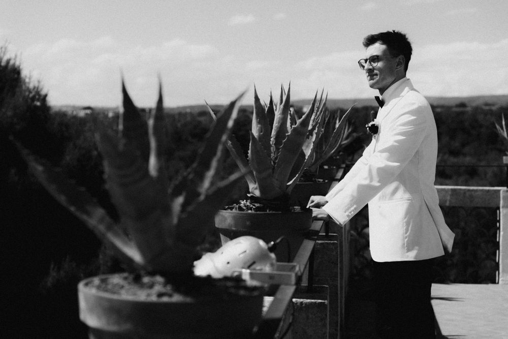 Black and white photo of a groom in a white tuxedo jacket standing beside potted succulents on an outdoor terrace.