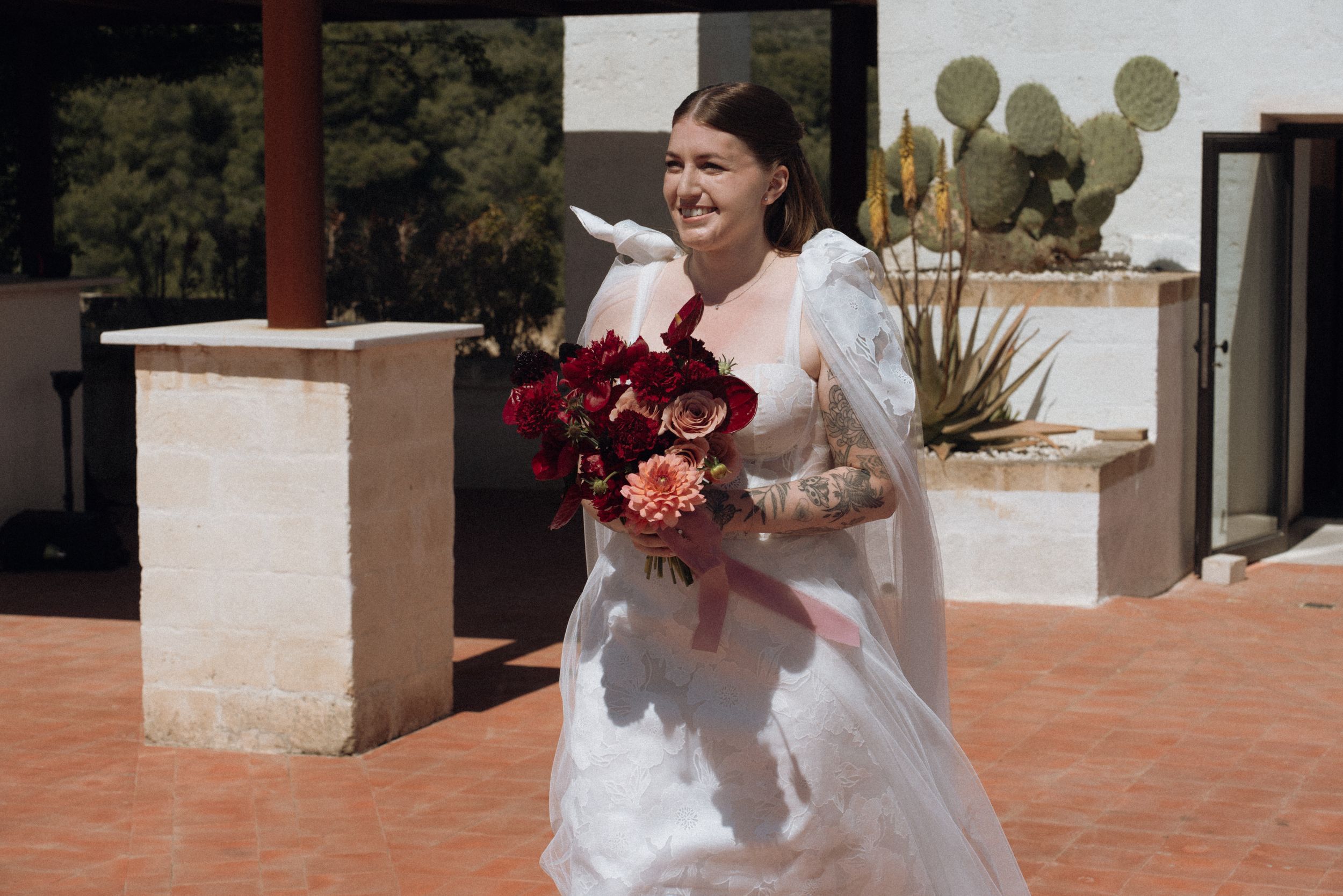 Smiling bride in white gown holding red and coral bouquet walks outdoors near adobe wall and cactus.