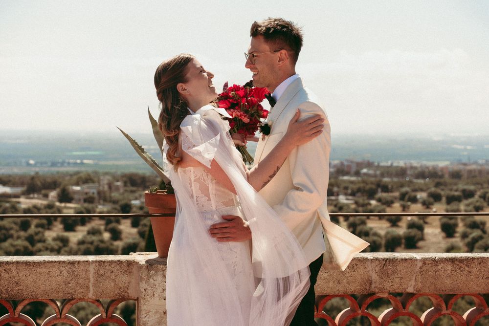 A bride and groom embrace on a scenic balcony, she holds red roses, wearing a white gown as he wears a cream suit.