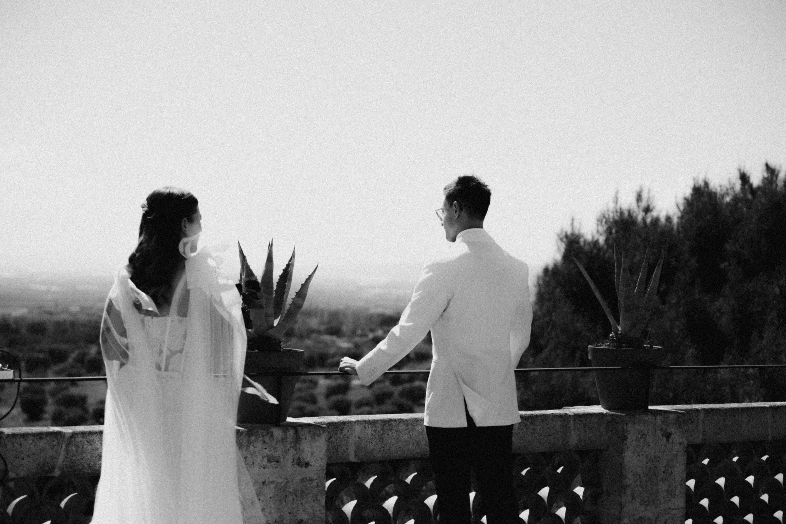 Black and white photo of a bride and groom holding hands on a balcony overlooking a scenic landscape.