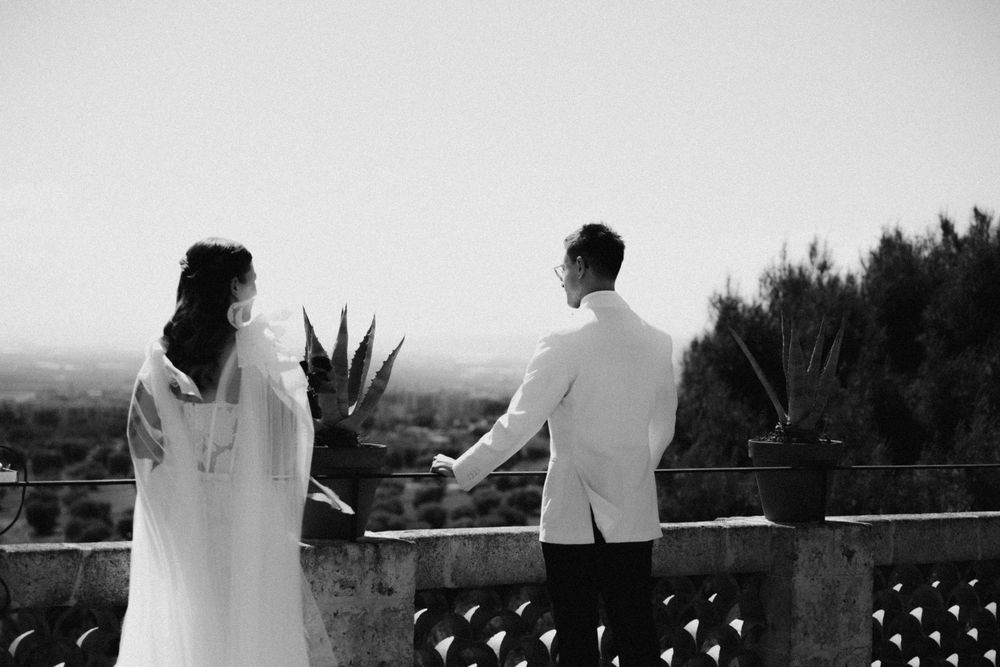 Black and white photo of a bride and groom holding hands on a balcony overlooking a scenic landscape.